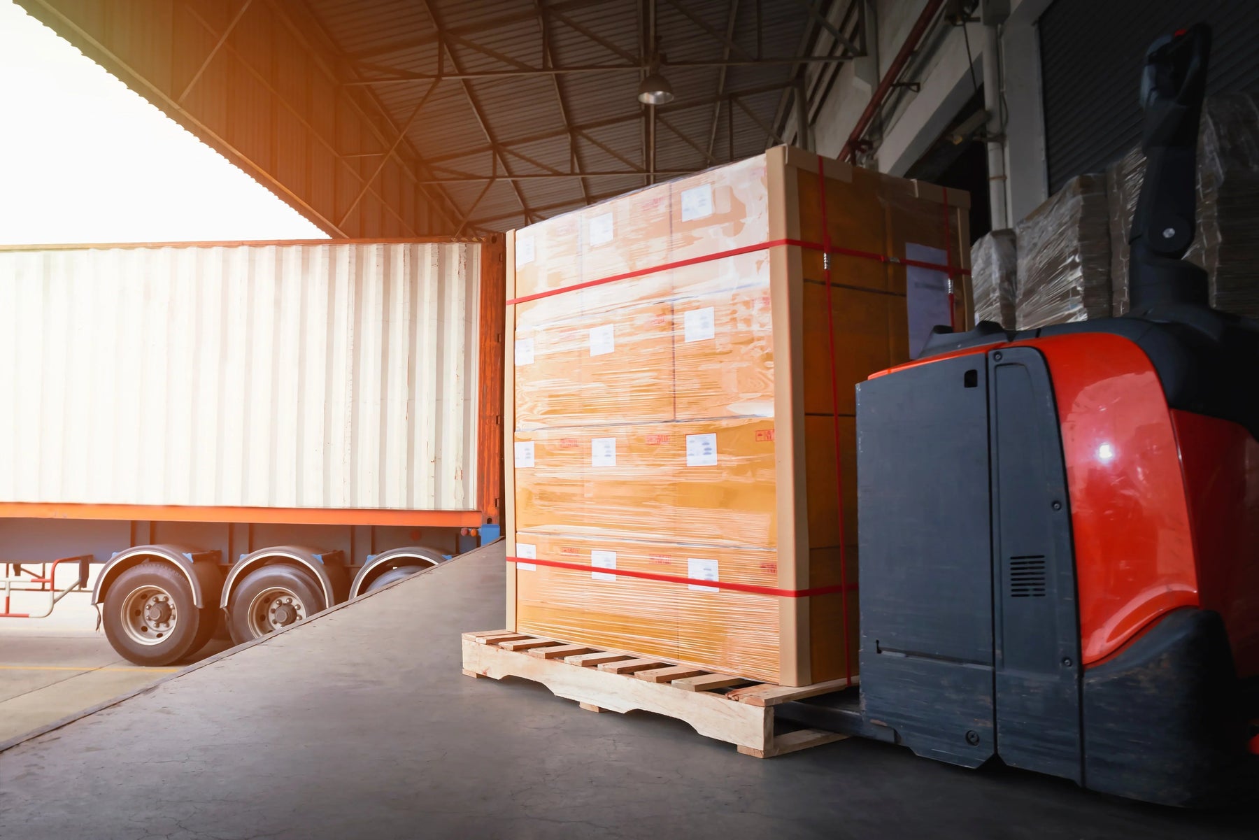 Electric pallet jack moving shrink-wrapped boxes onto a truck in a warehouse loading dock.