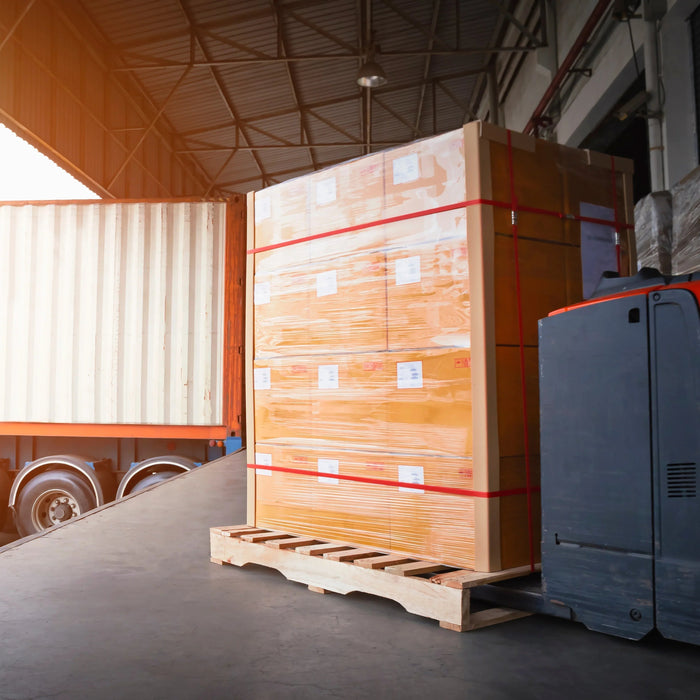 Electric pallet jack moving shrink-wrapped boxes onto a truck in a warehouse loading dock.