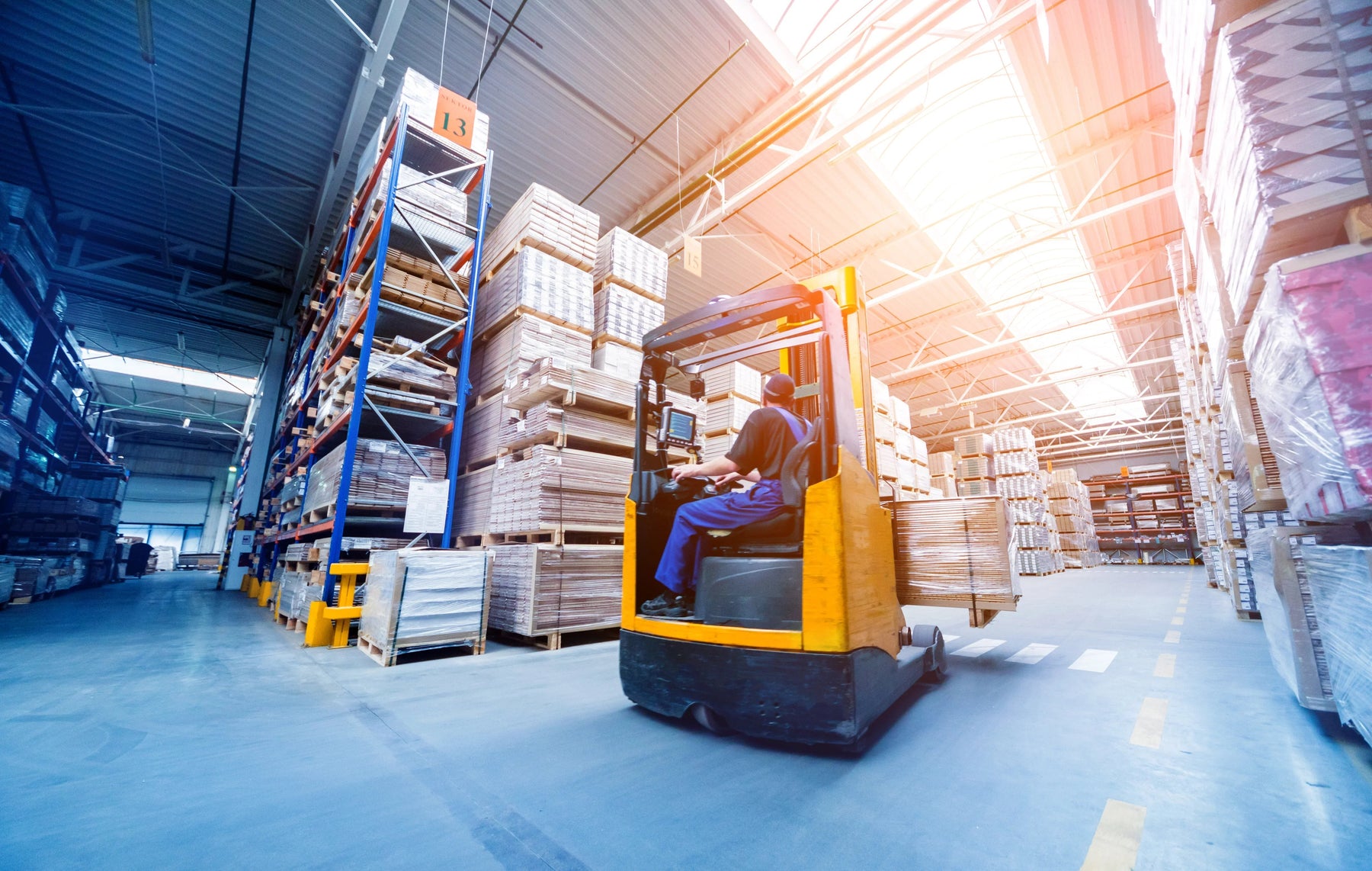 Worker driving forklift in warehouse aisle with stacked pallets and shelving