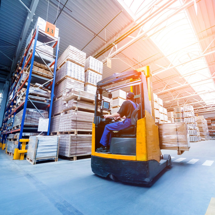 Worker driving forklift in warehouse aisle with stacked pallets and shelving